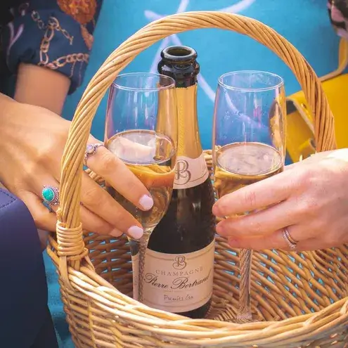 Two hands holding champagne glasses over a wicker basket containing a champagne bottle.