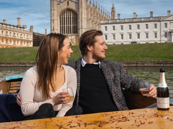 A young couple in a punting boat, holding glasses and laughing.