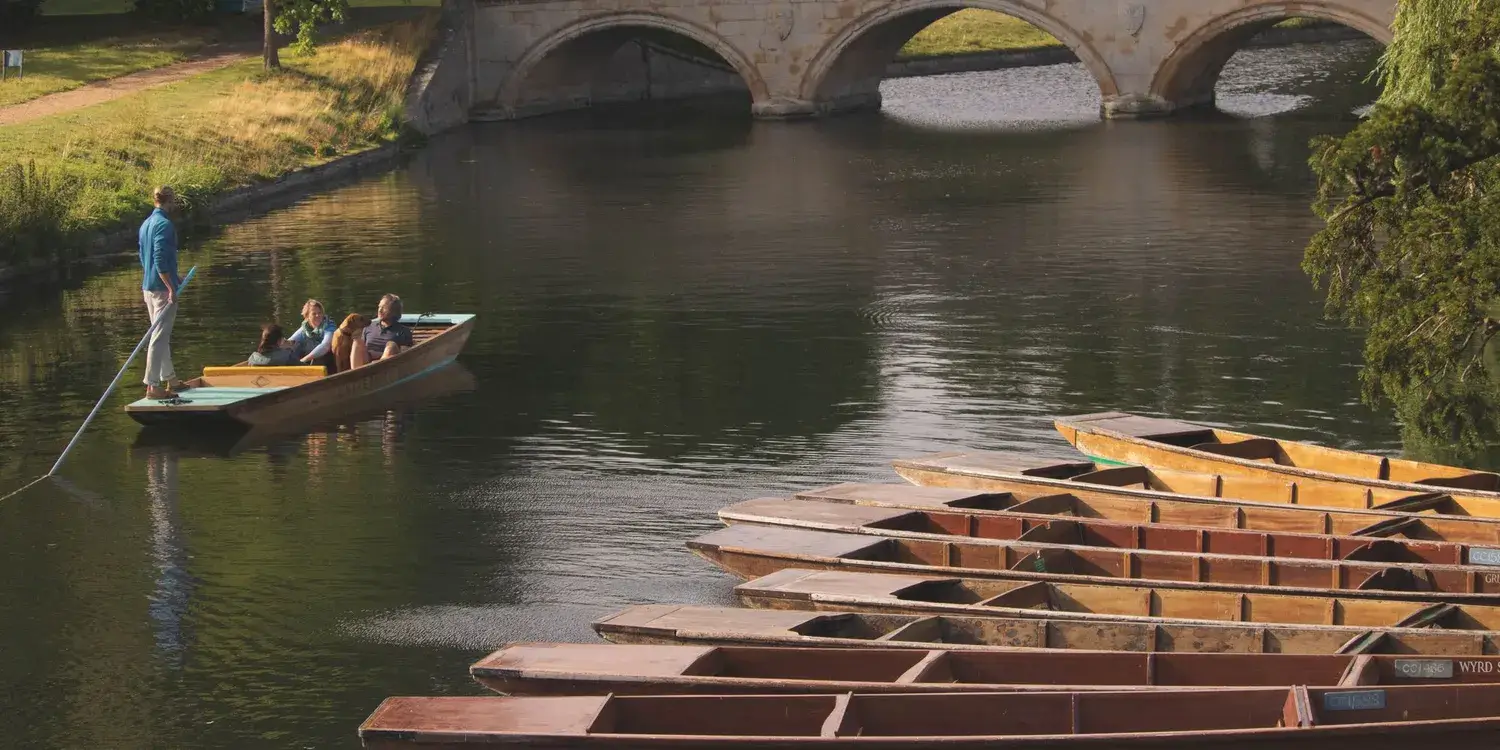 A wide shot of a party of people in a punting boat sailing past a group of empty, moored boats.