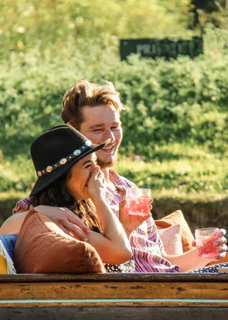 A young couple laughing and holding drinks in a punting boat.