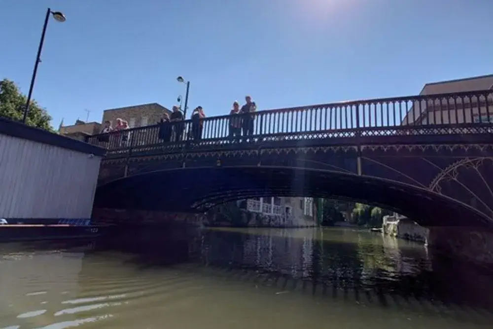 A view from the river of Magdalene Bridge, an intricately detailed bridge, with pedestrians looking over the railings at the boats below.