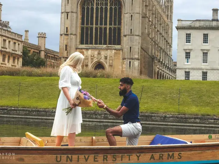 A man on one knee, proposing to a woman whilst in a punting boat in front of the King's College Chapel. The woman is wearing a white dress and holding a bouquet of flowers.