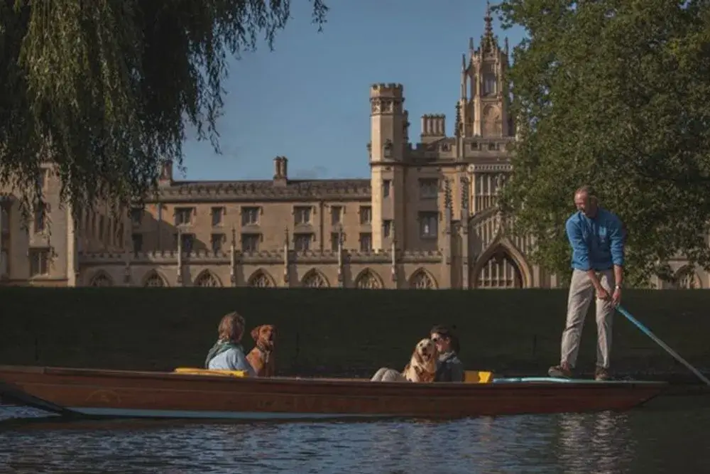 A couple of people and their dogs in a punting boat, in front of a grand, stone building, part of St John's New Court.