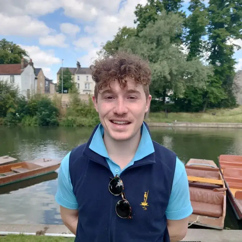 A photo of Tom, a young man with short, curly hair, smiling and standing in front of the river Cam.