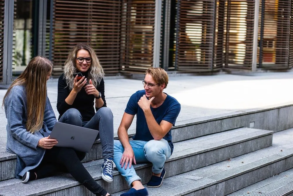 Three young adults sitting on steps outdoors, smiling and looking at a laptop.