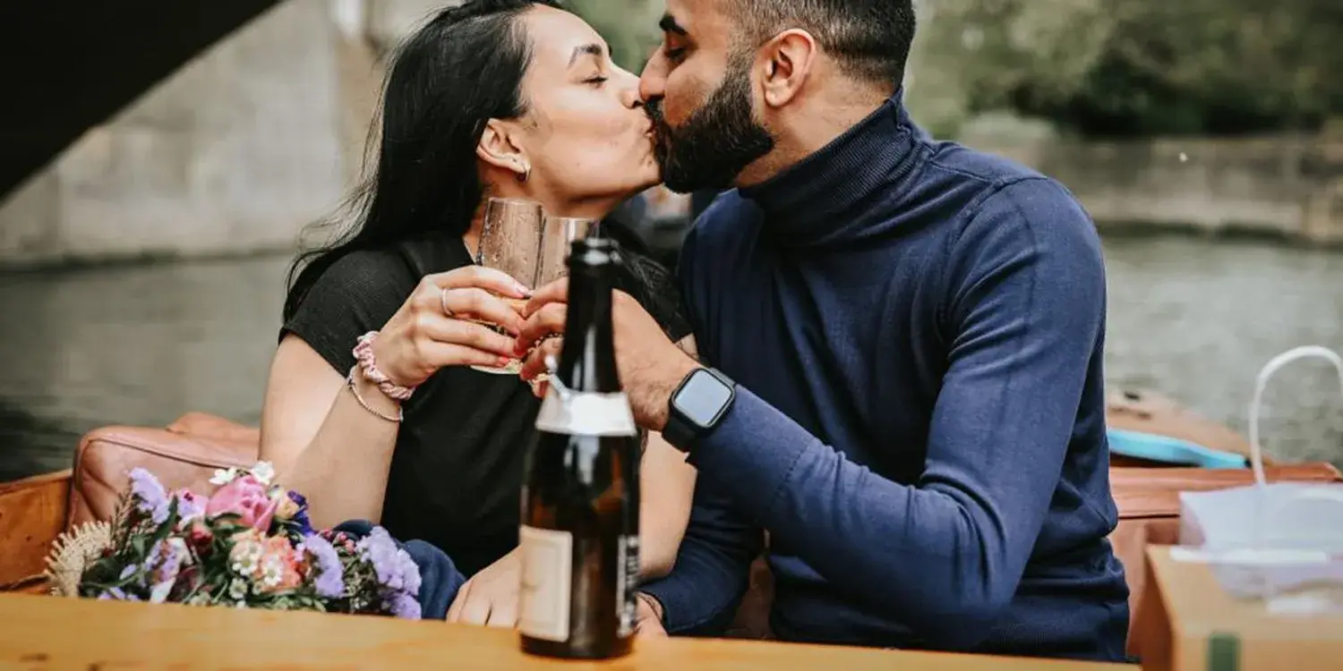 A young couple kissing and toasting glasses while sitting in a punting boat.