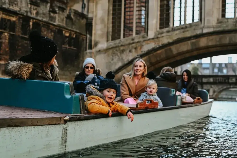 A family with children punting in Cambridge, passing beneath the famous Bridge of Sighs - gothic revival splendor. The children are leaning out of the boat and smiling.