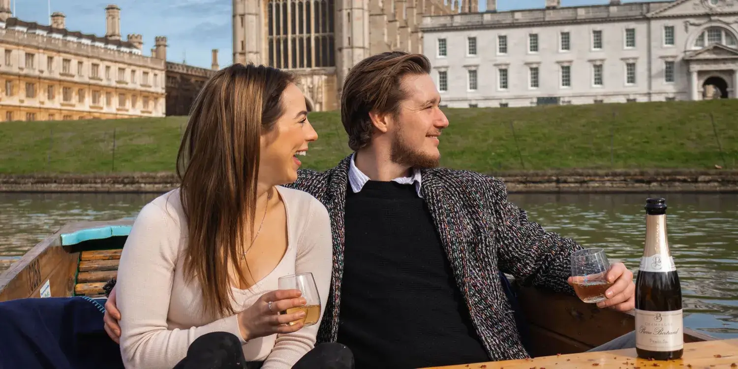 A young couple in a punting boat, holding glasses and laughing.