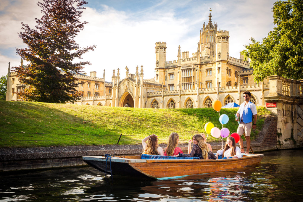 A party of people on a punting boat with colourful balloons and an impressive college building in the background.