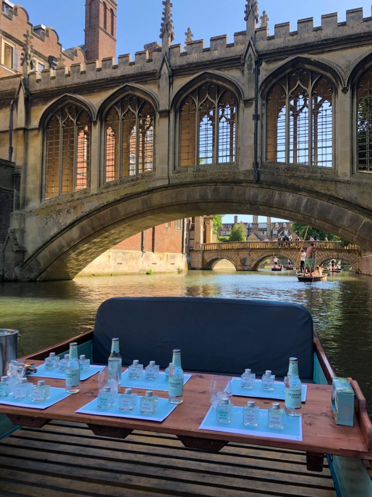 Six place settings consisting of the Cambridge Distillery Gin Trio laid out neatly on a punting boat with 2 impressive stone bridges in the background.
