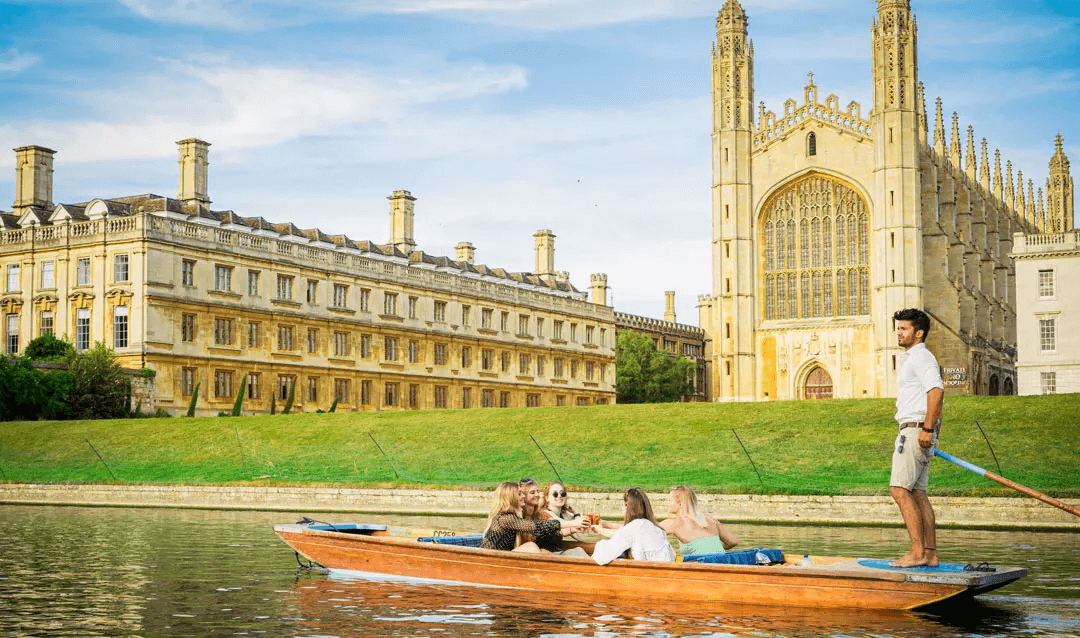 A traditional punt travels down the River Cam in Cambridge, past King's College Chapel on a beautiful sunny afternoon. A guide pushes a group of friend's enjoying a Pimm's as they glide.